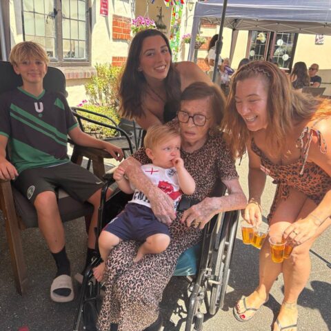 An elderly person in a wheelchair holding a child, surrounded by smiling family members outdoors.