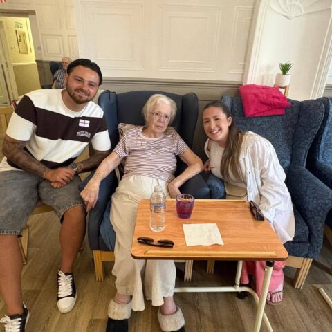 An elderly resident sitting with two visitors indoors, holding hands and enjoying a quiet moment together.