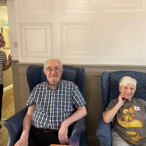 Two older adults seated in patterned armchairs in a cosy indoor lounge.