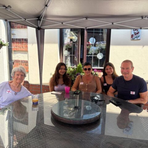 Five people sitting around an outdoor glass table under a canopy, wearing England flag stickers.