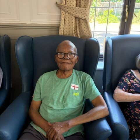 An older individual sitting in a blue armchair wearing a green shirt with an England flag emblem.