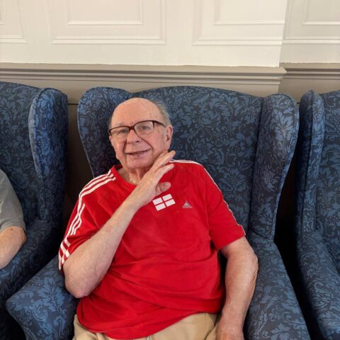 An older individual sitting in a blue armchair wearing a red shirt with an England flag emblem.