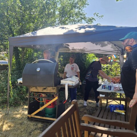 Four people preparing food at an outdoor BBQ under a canopy during a summer event.