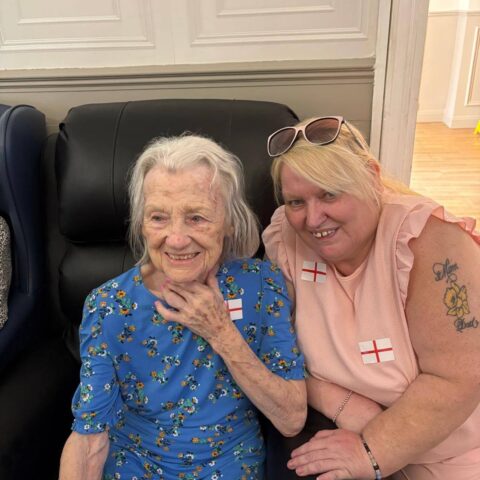 Two people sitting together indoors, one in a blue floral dress and the other in a pink outfit with England flag stickers.