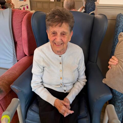 Smiling resident wearing a light blue cardigan seated in the lounge