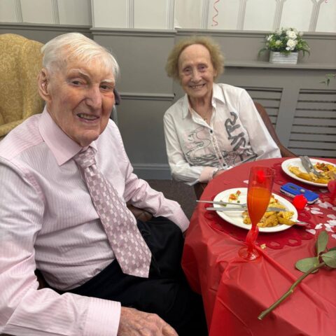 Couple seated together at a decorated anniversary table with a meal and rose