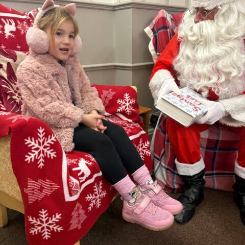 Child wearing a pink coat seated beside Santa on a decorated Christmas chair