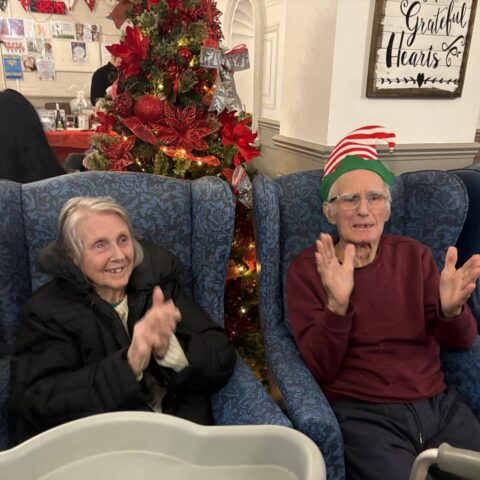 Two residents seated in armchairs clapping during festive entertainment