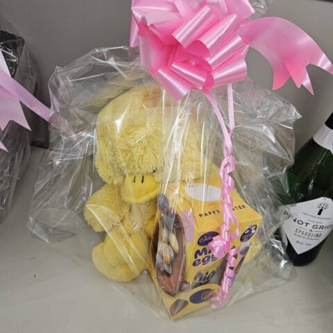 Cellophane-wrapped gift hamper containing a yellow teddy bear and treats with a pink bow