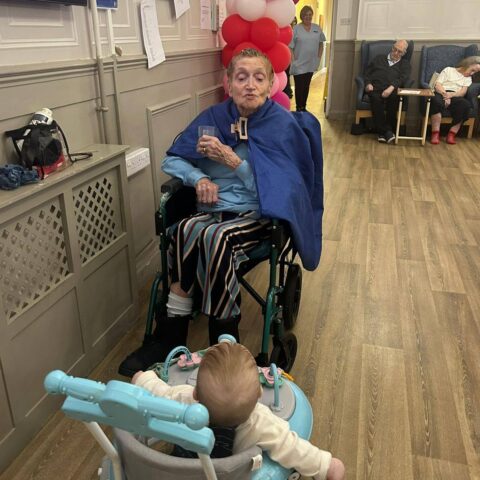 Resident smiling near a young child in a baby walker during a lounge visit