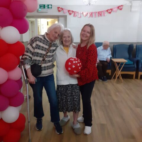 Three smiling people posing with a heart-patterned balloon beneath a pink and red balloon arch