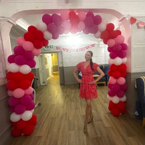 Performer in a red dress standing beneath a pink red and white balloon arch