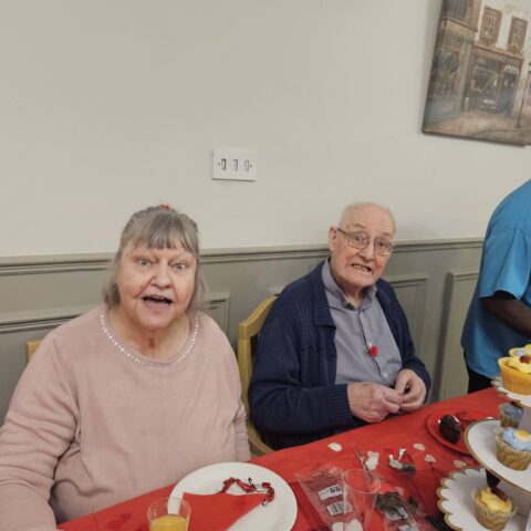 Two smiling residents seated beside a tiered cupcake stand at a red decorated table
