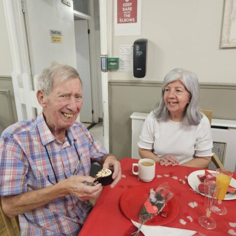 Smiling resident holding a cupcake while seated with a visitor at a red decorated table
