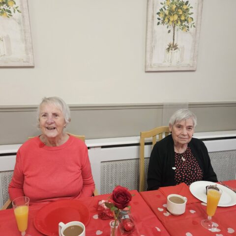 Two residents seated together at a red table with tea orange juice and a rose decoration