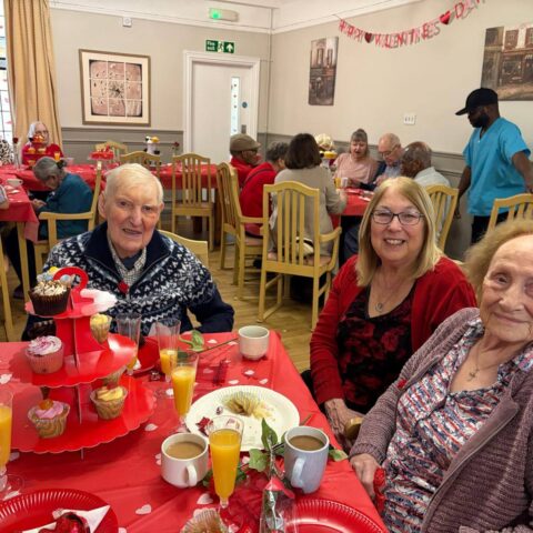 Three smiling residents seated at a red decorated table with cakes drinks and heart decorations