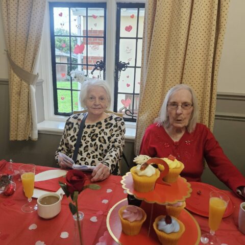 Two residents seated together at a red decorated table with cupcakes tea and orange juice