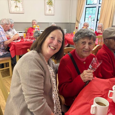 Smiling visitors and residents seated together around a red table with tea