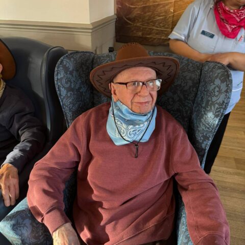 Resident smiling in the lounge while wearing a brown cowboy hat and neck scarf