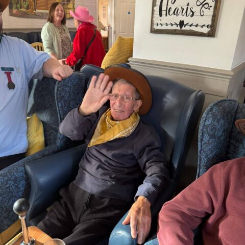 Resident wearing a cowboy hat and waving while seated in the lounge