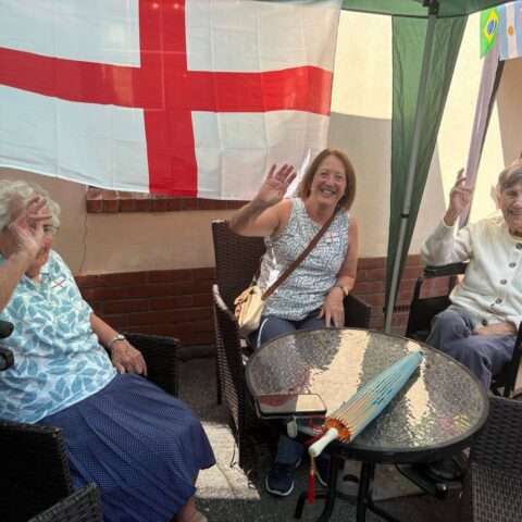 Residents sitting outdoors under a canopy decorated with international flags, raising their hands in celebration.