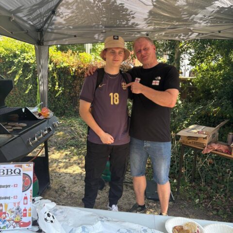 Two people standing at a summer BBQ wearing football-themed clothing, smiling beside a grill with burgers cooking.