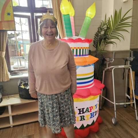 Resident wearing a gold crown beside a colourful inflatable birthday cake at Nightingale House